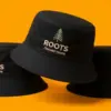 Три чёрные панамы ROOTS на оранжевом фоне, центральная — в повороте три четверти с вышивкой хвойного дерева на тулье