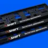 Три глянцевые рамки для номера SHIFT уложены каскадом под углом, оранжевый логотип контрастирует с чёрным пластиком