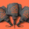 Три складных зонта Aura веером на коралловом фоне — крупные оранжевые логотипы на тёмно-серых куполах