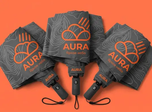 Три складных зонта Aura веером на коралловом фоне — крупные оранжевые логотипы на тёмно-серых куполах